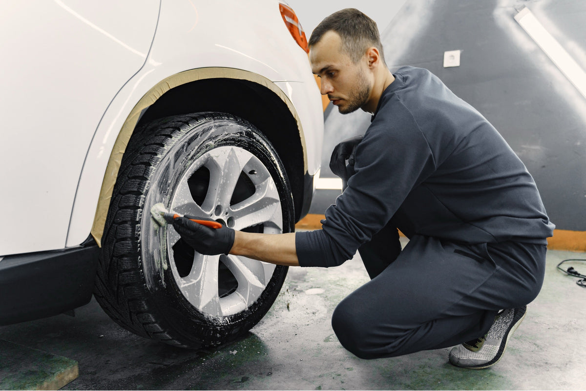 A detailer kneeling beside a white SUV, scrubbing a foam-covered alloy wheel with a detailing brush