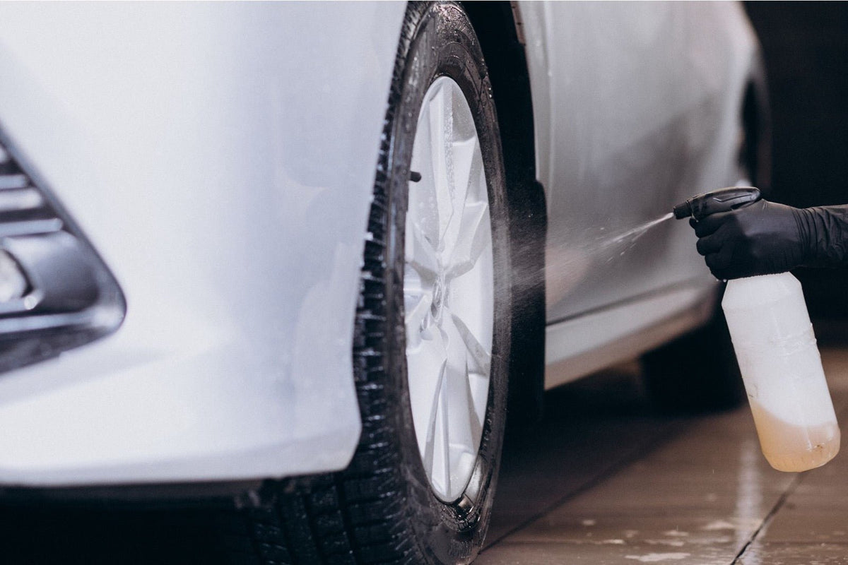 A gloved hand sprays cleaner onto the wheel of a white car at a detailing station