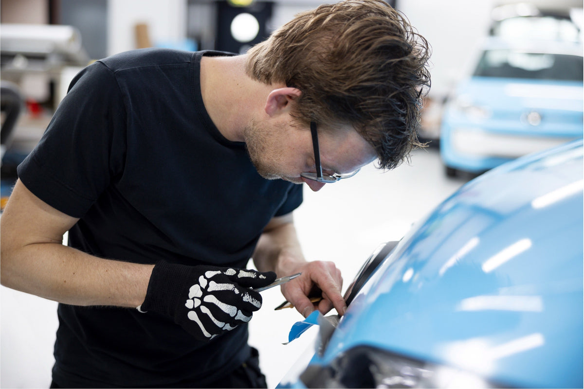 Detailer in a black shirt and gloves inspecting the front fender of a light blue car in a workshop.