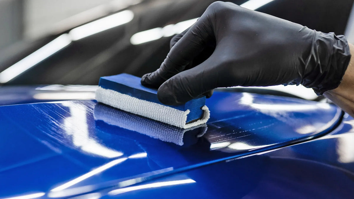 Gloved hand applying ceramic coating with microfiber applicator block to glossy blue car hood