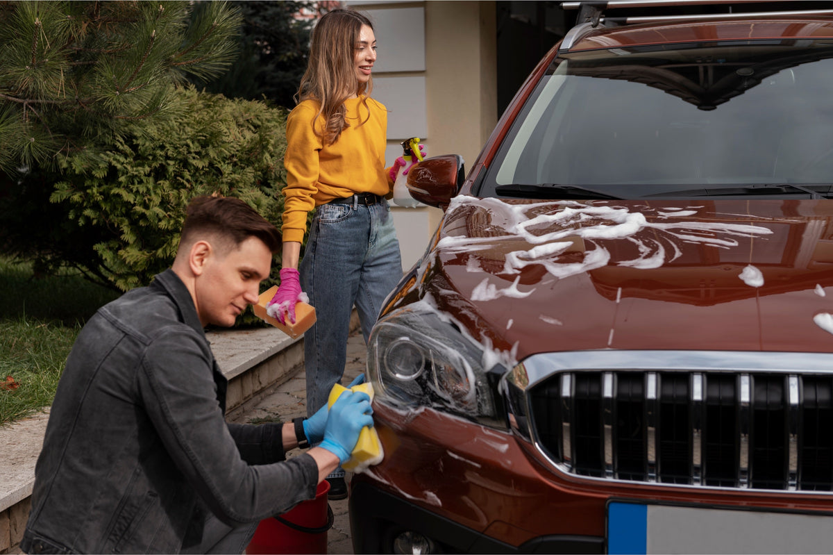 Man and woman detailing and hand-washing a red SUV with sponges and foam in a driveway.