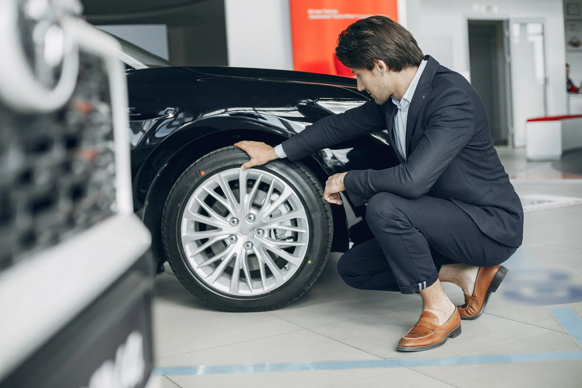 Man in a navy suit crouching beside a black car to inspect its silver alloy wheel and tire