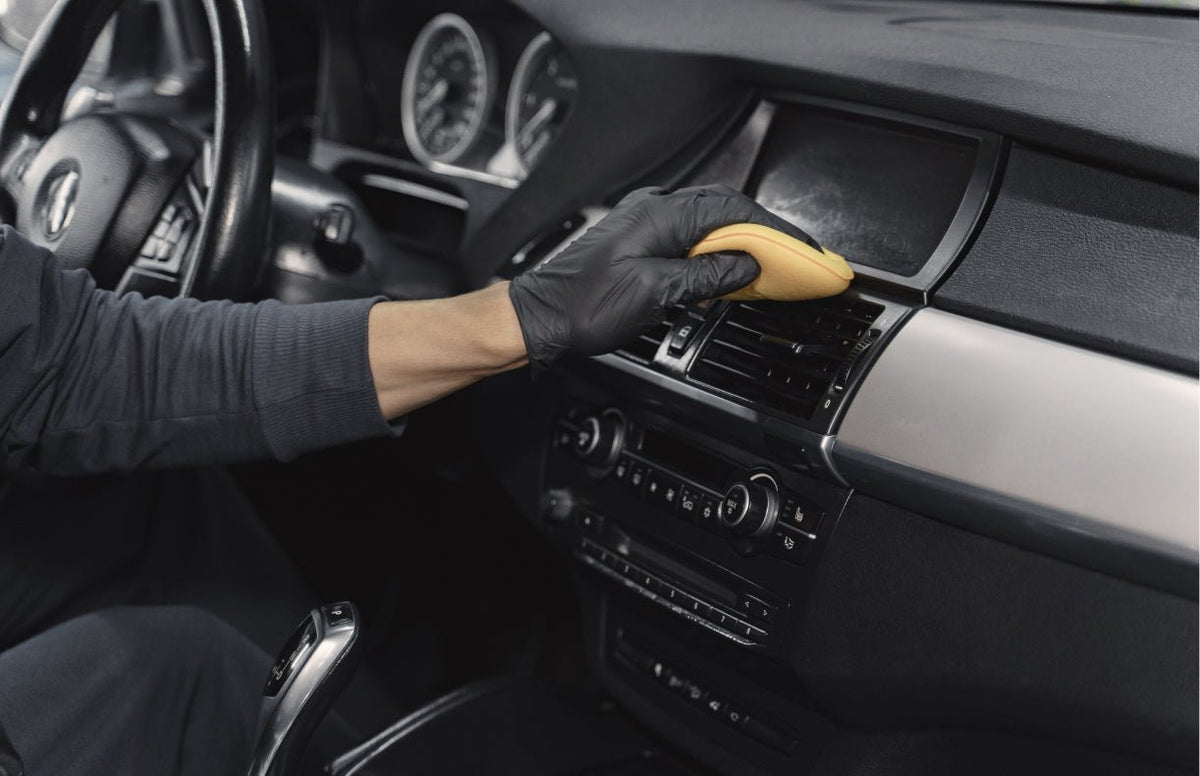 A gloved hand wiping a car dashboard and plastic trim with a yellow microfiber applicator.