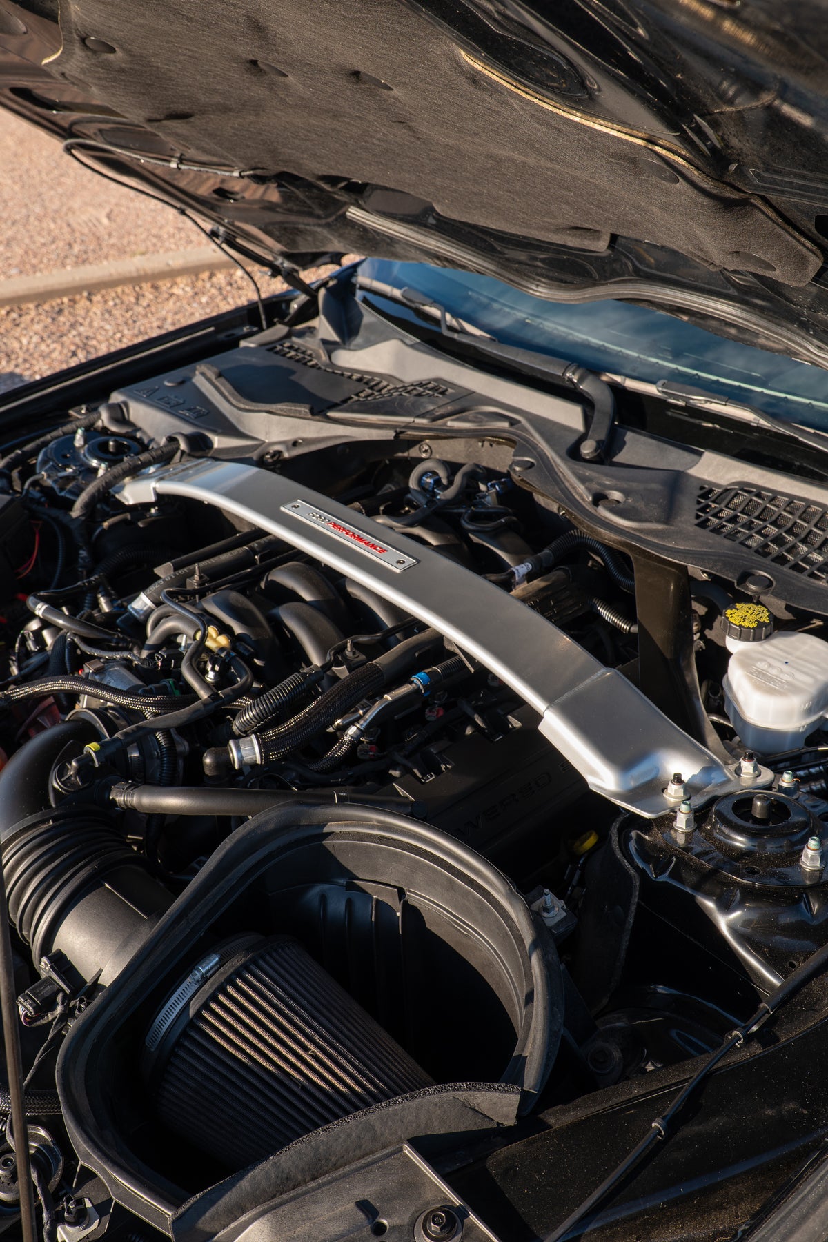 A close-up view of a car engine bay with a polished silver engine cover featuring a red and black logo, alongside a black air filter and various engine components.