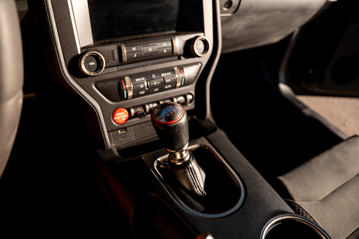 Close-up of a manual gear shift knob with a black leather-wrapped handle and red and blue patterned top, set in a dark car console.