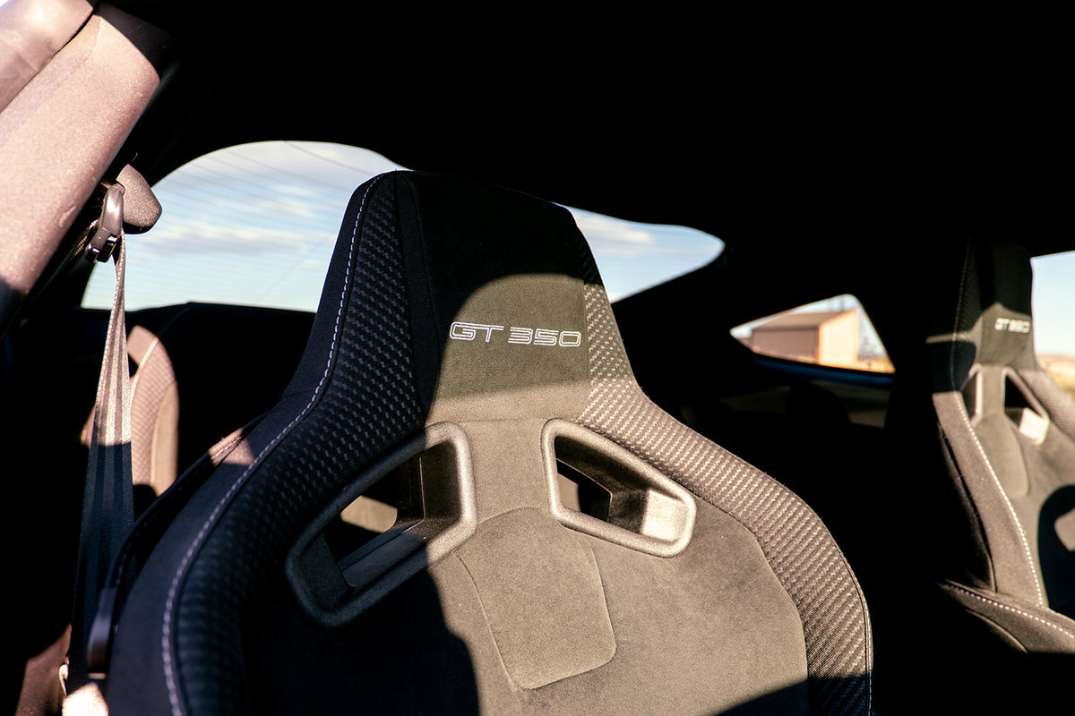 Close-up of a car interior showing a black and gray racing bucket seat with 'GT 350' embroidered on the headrest, with another matching seat partially visible on the right.