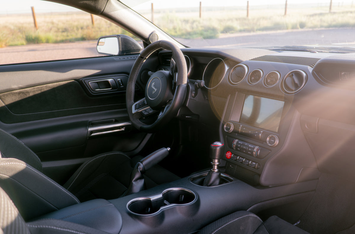 A close-up view of a car's interior dashboard, steering wheel, center console, and gear shift selector, photographed from the driver's side with bright natural light.