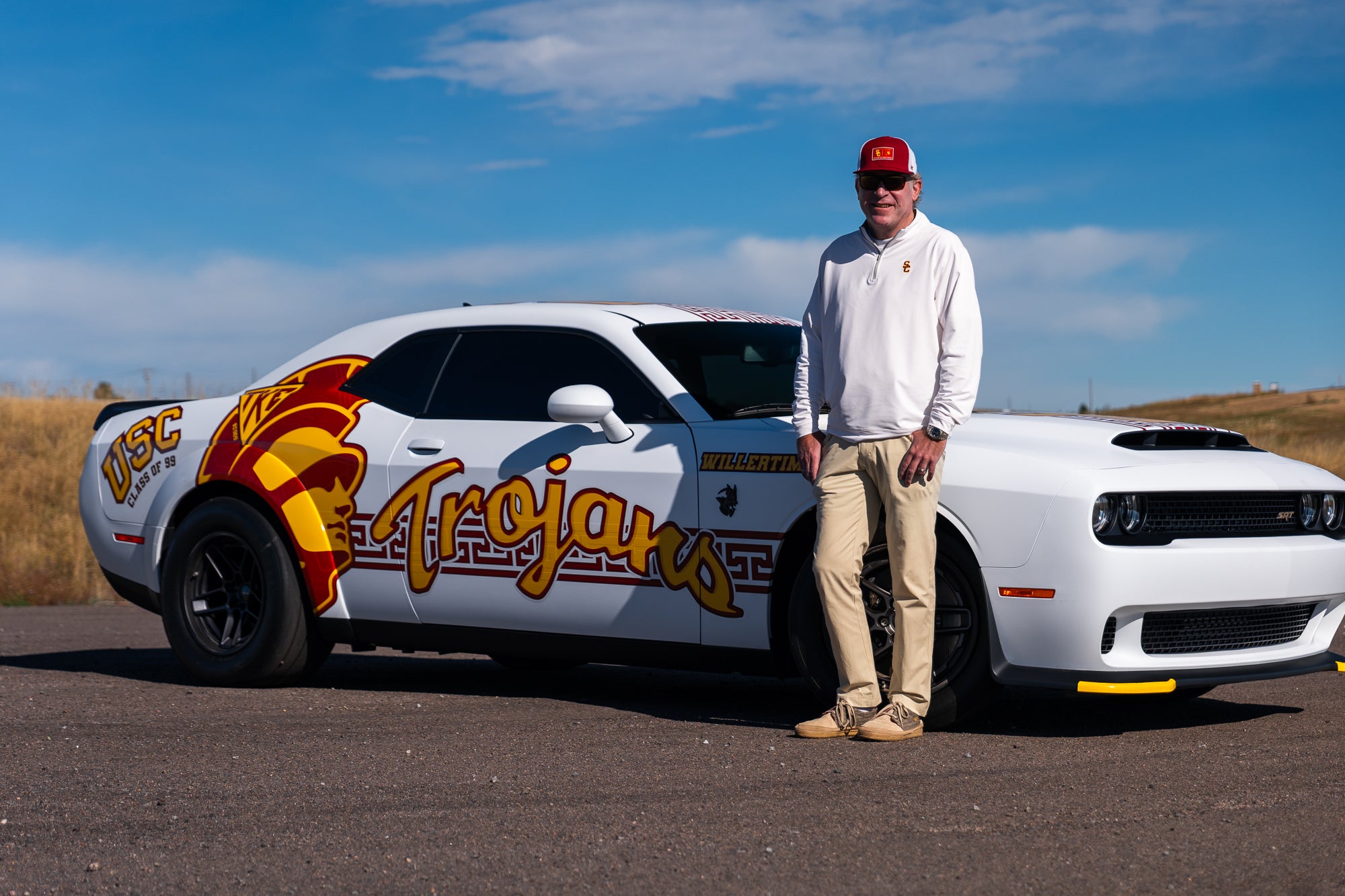 Man standing next to a white car with 'Trojans' branding on a road with a blue sky.